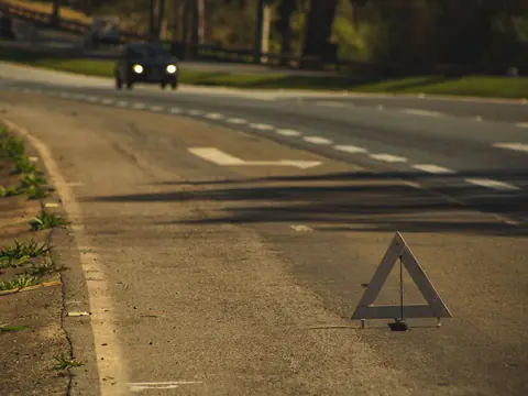 A reflective warning triangle standing on the edge of a road at dusk, with one car approaching in the distance