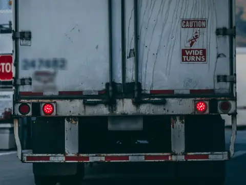 The rear doors of a white semitrailer with its rear impact guard, red and white reflective tape, red lamps and a Caution Wide Turns sign