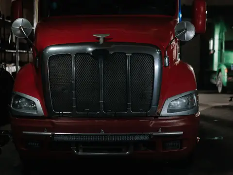 The front of a red semi truck cab with its chrome grille, headlights and mirrors, parked in a dim truck garage