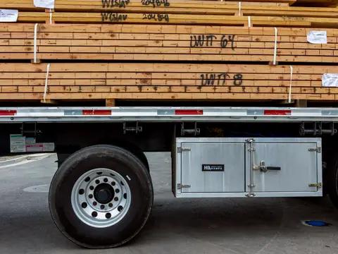 An orange semi truck hauling a flatbed trailer stacked with concrete blocks along a tree-lined road