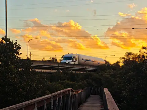 A tank truck crossing a raised highway overpass against an orange sunset sky, seen past trees and a footbridge
